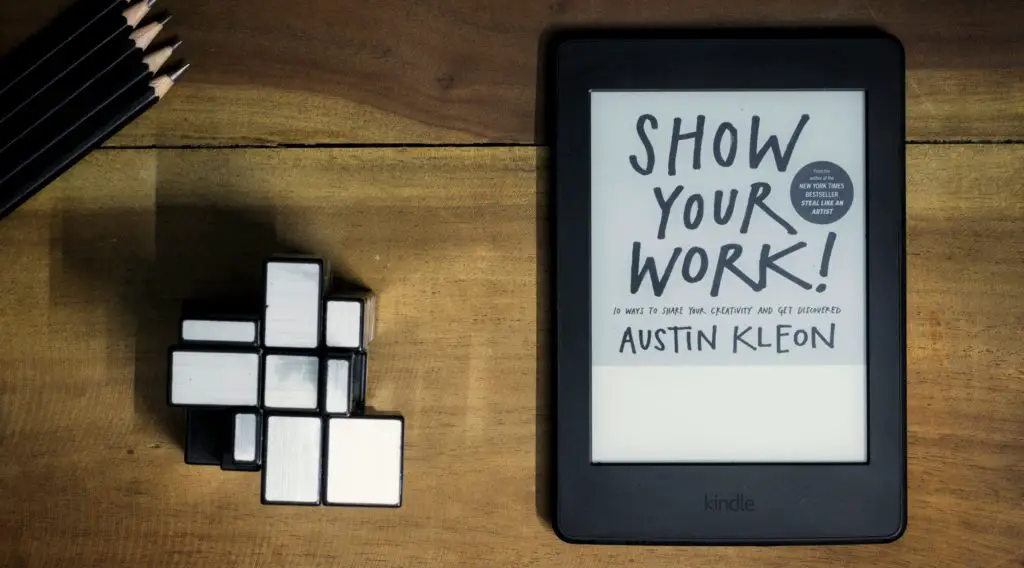 Kindle on a wooden desk with an ebook open. A Mirror- Rubik's cube and some pencils are also on the desk.